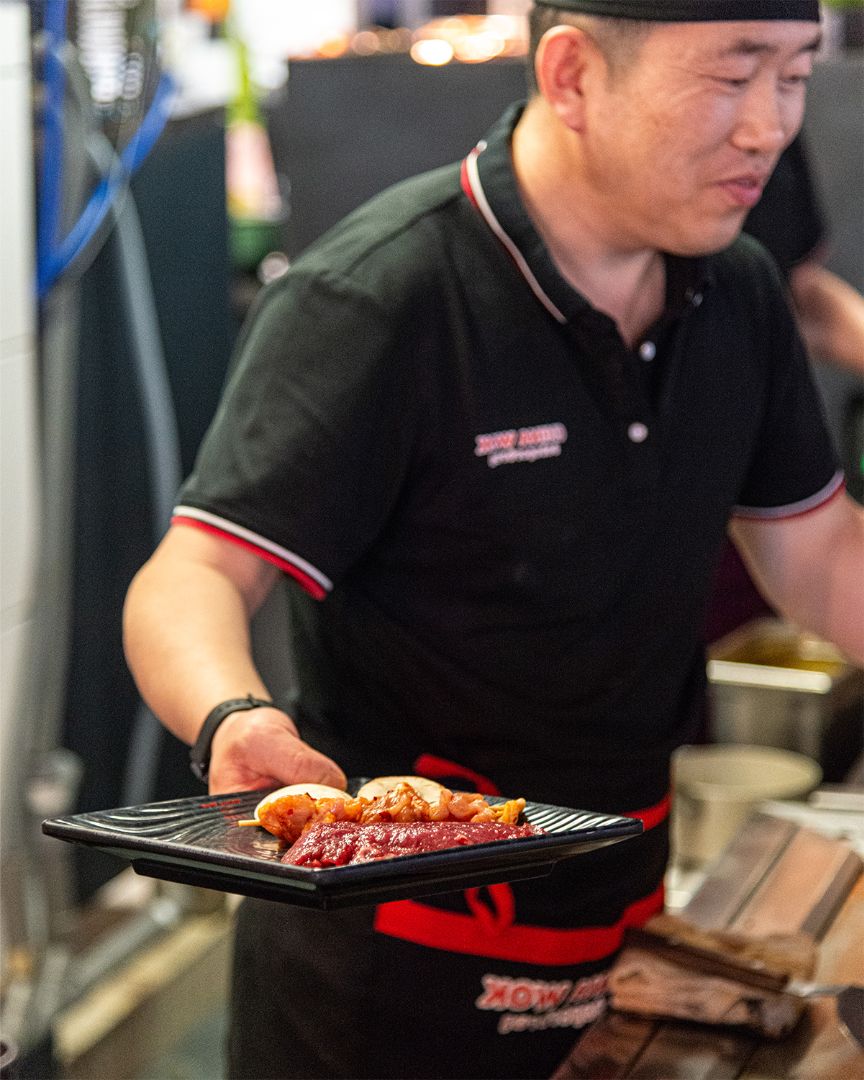 Een kok in uniform van China Wok houdt een zwart bord vast met rauwe gemarineerde vleesgerechten, klaar om gegrild te worden in een open keuken.